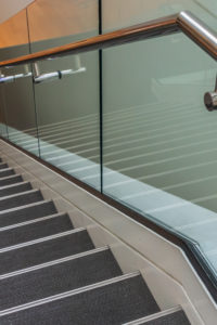 Stairs are leading downward to a small platform. The stair steps are gray. A metal handrail and glass panels are next to the stairs. Stairs are leading downward to a small platform. The stair steps are gray. A metal handrail and glass panels are next to the stairs.